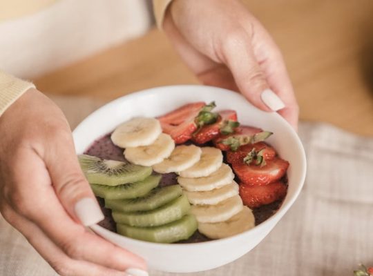Delicious smoothie bowl topped with fresh fruits like kiwi, banana, and strawberries, held by manicured hands.