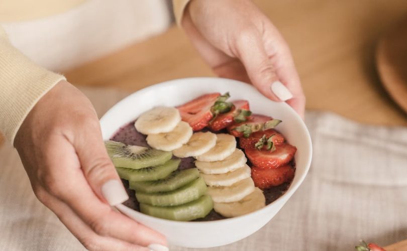 Delicious smoothie bowl topped with fresh fruits like kiwi, banana, and strawberries, held by manicured hands.